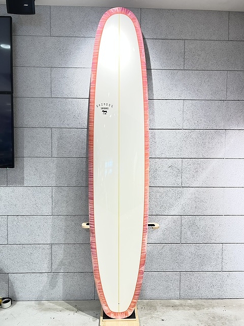 White surfboard with a pink border standing upright on a wooden rack against a gray tiled wall in an indoor display.