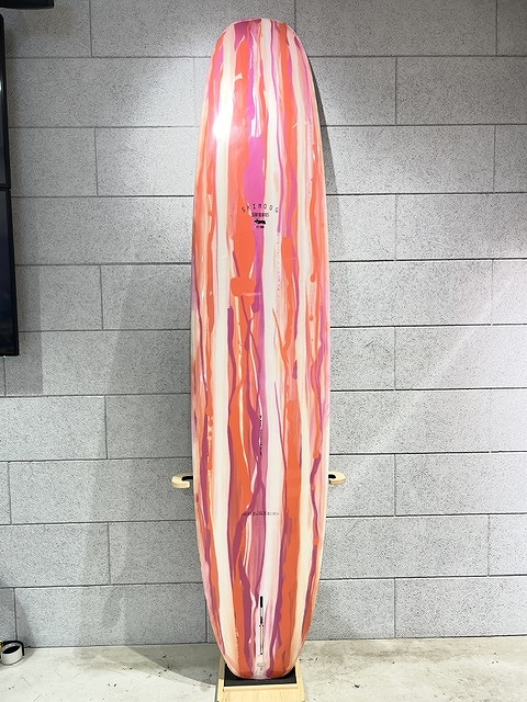 Pink, orange, and white striped surfboard standing upright against a gray brick wall on a wooden stand.