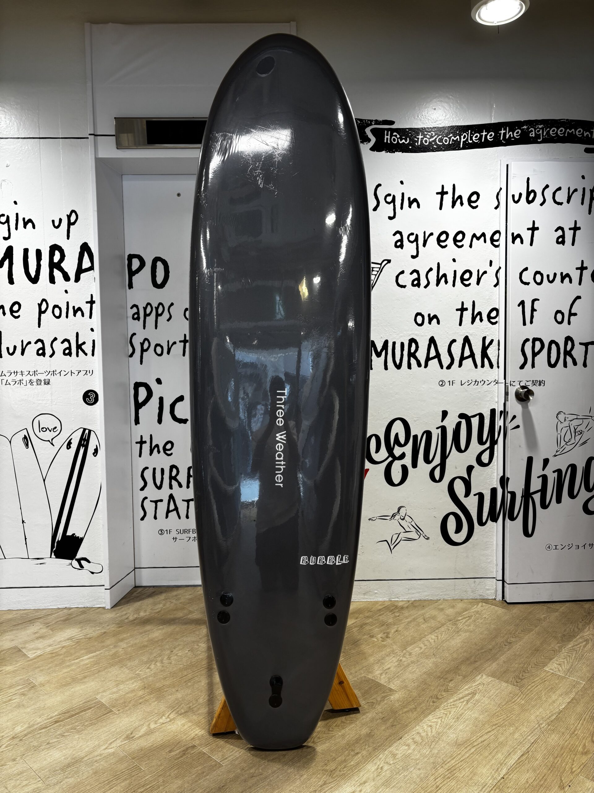 IMG_0506 - SHOP NEWS | ムラサキスポーツオンラインストア 通販 Glossy black surfboard standing upright on a wooden rack indoors, with white walls covered in bold, hand-written surfing text in the background.