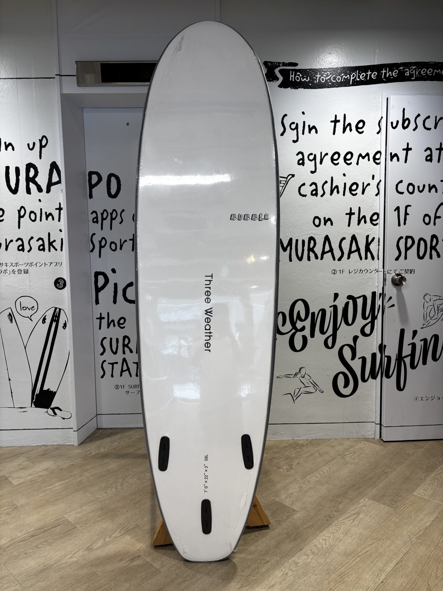 IMG_0507 - SHOP NEWS | ムラサキスポーツオンラインストア 通販 White surfboard standing upright indoors on a wooden floor, with black graffiti-style text and drawings on the walls behind it, and black fin slots visible at the bottom of the board.