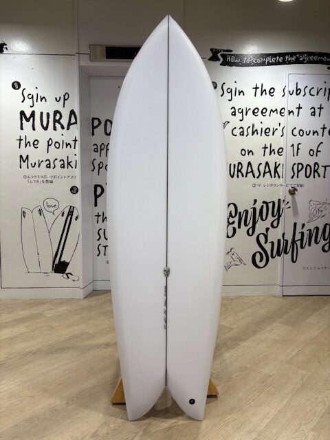 IMG_7755 - SHOP NEWS | ムラサキスポーツオンラインストア 通販 White surfboard standing upright on a wooden stand inside a shop, with bold black handwriting murals on the walls around it.