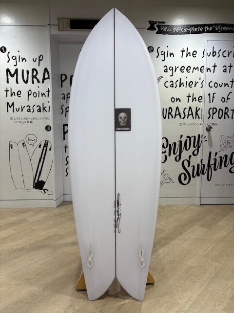 IMG_7756 - SHOP NEWS | ムラサキスポーツオンラインストア 通販 Two white surfboards standing upright on a wooden rack inside a surf shop, with black surfing-themed wall art; center board has a small skull sticker.