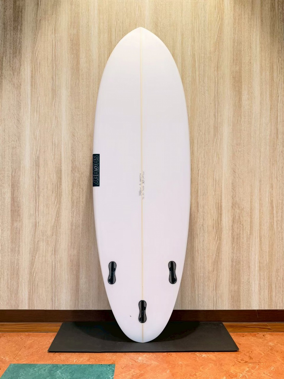 LINE_ALBUM_2026 フリーダム_260601_2 - SHOP NEWS | ムラサキスポーツオンラインストア 通販 White surfboard standing upright against a wood wall on a black mat, three fin boxes visible on the bottom half.